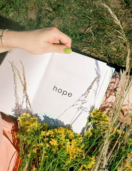 Hand holding an open book displaying 'hope' among yellow flowers in a field.